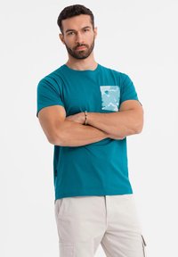 Teal cotton t-shirt with short sleeves, featuring a camouflage-patterned pocket. Smooth texture, casual design, with a round neckline.