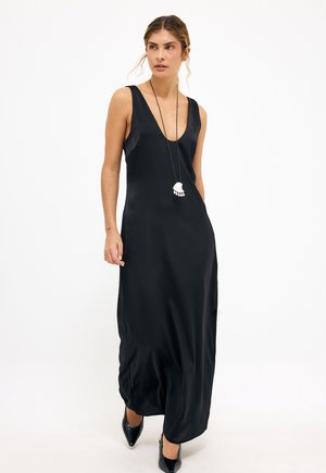 Kobieta w czarnej, bezrękawnikowej sukni maxi z głębokim dekoltem w serek, czarnych, spiczastych szpilkach, długim naszyjniku z zawieszką i włosami związanymi do tyłu.