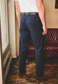 Laadukkaat tummansiniset housut, joissa on slim-fit-leikkaus, tekstuuri ja ruskea nahkavyö. Takana näkyy kaksi nappikiinnityksellä varustettua taskua.
