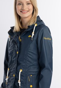 Jachetă impermeabilă bleu marin cu glugă, accente galbene, șnururi elastice, fermoar frontal și capse. Prezintă un logo pe piept.
