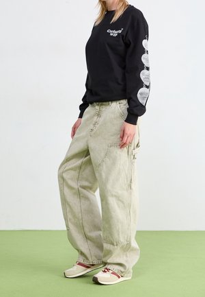 Osoba ubrana w czarną koszulę Carhartt WIP z długim rękawem ozdobioną srebrnymi wzorami serca na rękawie, szerokie beżowe spodnie i kremowe tenisówki z czerwonymi akcentami.