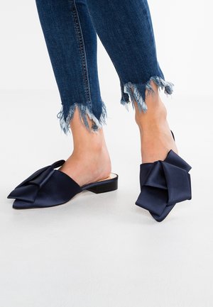 Granatowe satynowe sandały na obcasie typu slingback z dużą kokardą i małym, szerokim obcasem, zestawione z ciemnymi dżinsami o postrzępionych nogawkach.