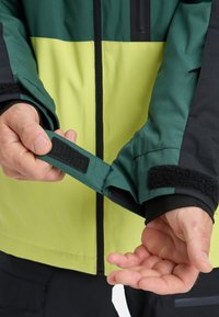 Green and black jacket with a yellow panel, featuring adjustable cuff with Velcro, and a smooth texture. Hand showing the cuff detail.