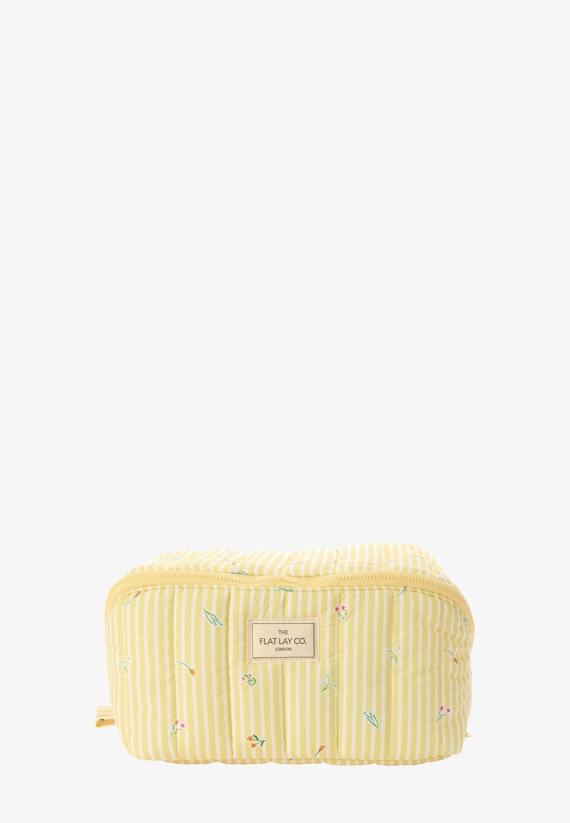 Pochette matelassée rayée jaune et blanche avec petits motifs floraux, forme rectangulaire, fermeture éclair, et étiquette "The Flat Lay Co. London" sur le devant.