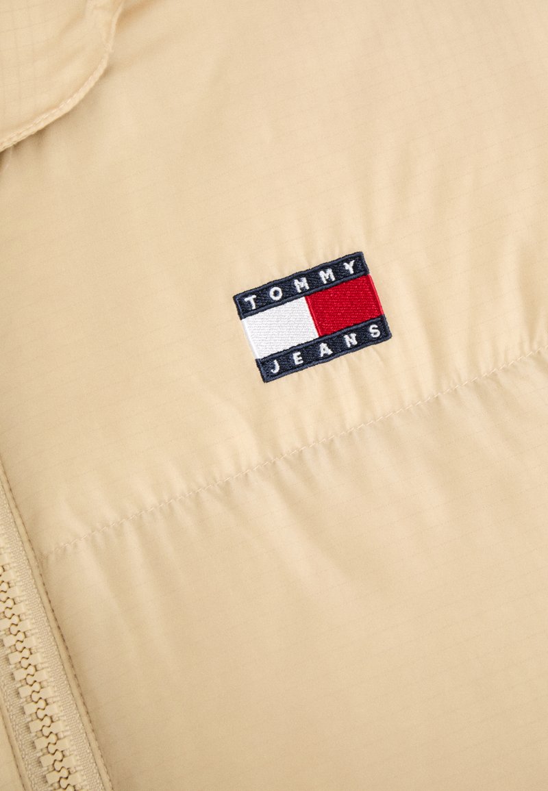 Beige gepolsterte Jacke mit Reißverschluss und einem gestickten rechteckigen Logo mit rot, weiß und blau Akzenten. Glattes, leichtes Material.