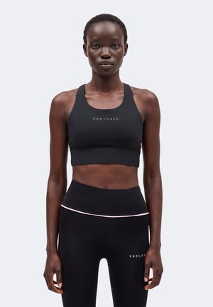 Femme portant un soutien-gorge de sport noir et des leggings avec "ENDLESS" imprimé, debout face à l'avant sur un fond clair uni.