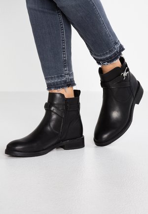 Sorte læderankelstøvler med sidelukning og dekorativ rem, båret med mørkegrå skinny jeans med frynset kant på lys gulvbaggrund.