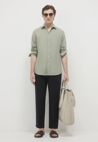 Uomo con camicia verde chiaro a bottone, pantaloni neri, sandali scuri, occhiali da sole, che tiene una borsa tote beige, in piedi di fronte a uno sfondo bianco semplice.