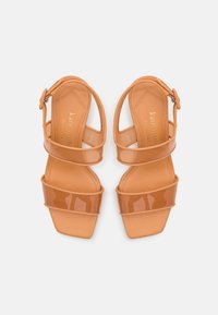 Tan patent leather sandals with a square toe, featuring three horizontal straps and a buckle on the ankle strap. Smooth texture and glossy finish.