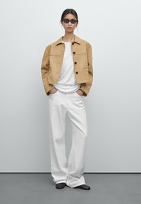 Tan suède cropped jasje met vier zwarte knopen, gedragen over een witte shirt. Gecombineerd met brede witte broeken en zwarte puntige schoenen. Stijlvolle zonnebril.