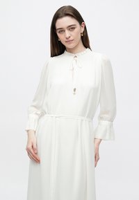 Bílé dlouhé šaty s volánovým límečkem, zavazovacím výstřihem a zvonovými rukávy. Hladká textura s úzkým pásem v pase.