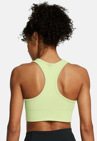Mujer con cabello rizado lleva un sujetador deportivo verde claro y leggings negros, vista por detrás contra un fondo blanco.