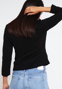 Haut noir texturé à manches longues avec un design court, associé à un jean en denim bleu clair. Met l'accent sur la vue de dos et le détail de l'épaule.