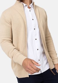 Cardigan beige en maille avec un col montant, une fermeture éclair à l'avant et des poignets côtelés. Porté par-dessus une chemise blanche à boutons marron.