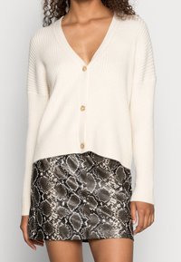 Cremfarbener strukturierter Cardigan mit V-Ausschnitt und Knöpfen, kombiniert mit einem Rock im Schwarz-Weiß-Pythonmuster, der eine figurnahe Silhouette zeigt.