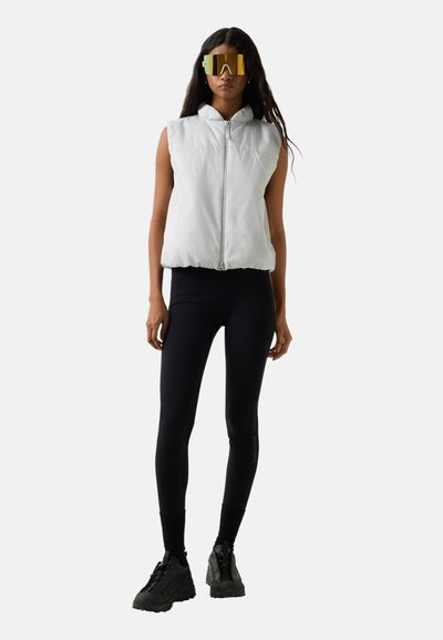 Kobieta ubrana w bezrękawnik biały puchowy, czarne legginsy, masywne czarne sneakersy oraz odbijające światło złote okulary przeciwsłoneczne typu wraparound, stoi na tle jednolitego tła.