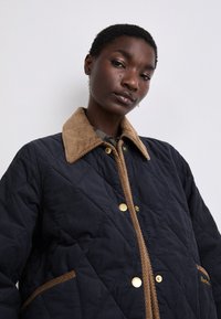 Casaco acolchoado azul marinho com colarinho de veludo cotelê bege, botões de pressão dourados e detalhes em corda castanha nos bolsos. Tecido texturizado com padrão em diamond.