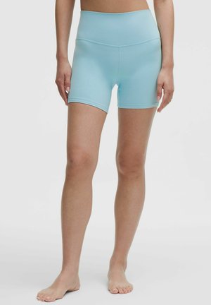 Vrouw draagt lichtblauwe, hooggetailleerde, aansluitende sportshorts en staat blootsvoets tegen een effen achtergrond.