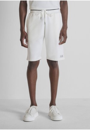 Uomo che indossa pantaloncini bianchi con coulisse e scarpe da ginnastica bianche, in piedi davanti a uno sfondo grigio chiaro uniforme.