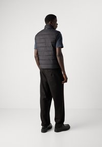 Padded black gilet with a high collar and sleeveless design worn over a short-sleeve navy shirt, paired with black trousers and shoes.
