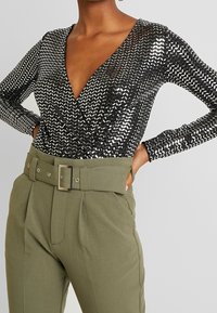 Schwarzer Pailletten-Body mit tiefem V-Ausschnitt und langen Ärmeln, kombiniert mit hoch taillierten olivgrünen Hosen, die einen breiten Gürtel mit silberner Schnalle haben.