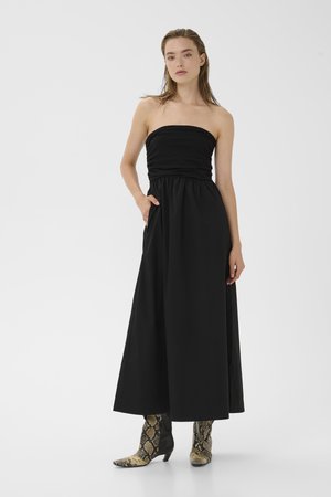 Kvinde iført sort strapless maxikjole med lommer og ankelstøvler med slangeprint, stående foran en ensfarvet hvid baggrund.