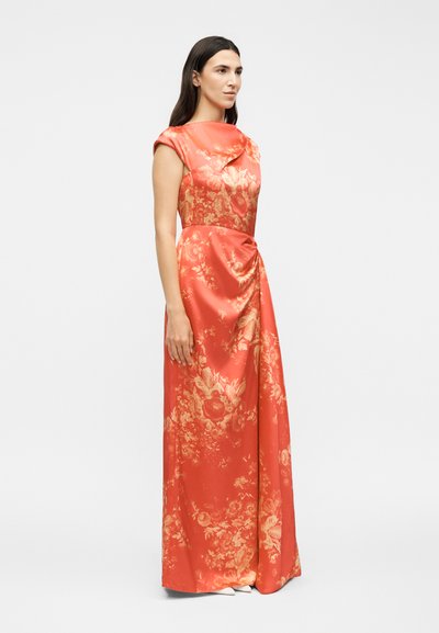 Γυναίκα στέκεται φορώντας ένα κοραλλένιο φόρεμα μέχρι το πάτωμα χωρίς μανίκια με χρυσά λουλουδένια σχέδια και μια ρυτιδιασμένη λεπτομέρεια στη μέση.