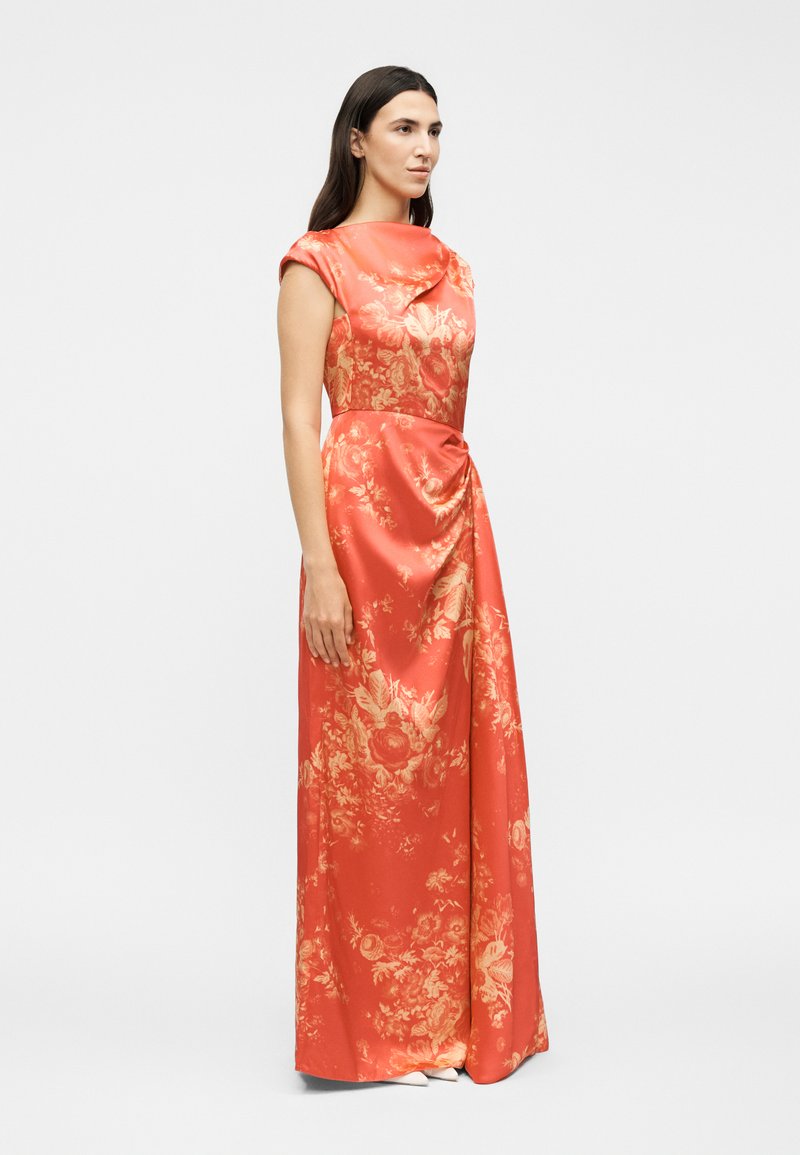 Woman standing wearing a floor-length sleeveless coral dress with gold floral patterns and a gathered detail at the waist.