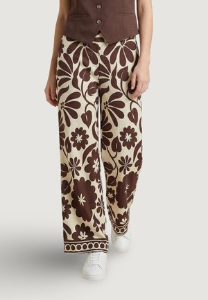 Pantaloni crema a gamba larga con grande stampa floreale marrone indossati con gilet marrone abbottonato e sneakers bianchi su sfondo neutro.