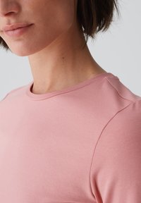 Hellrosa T-Shirt mit Rundhalsausschnitt, kurzen Ärmeln und glatter Textur, das subtile Nähdetails an der Schulternaht aufweist.