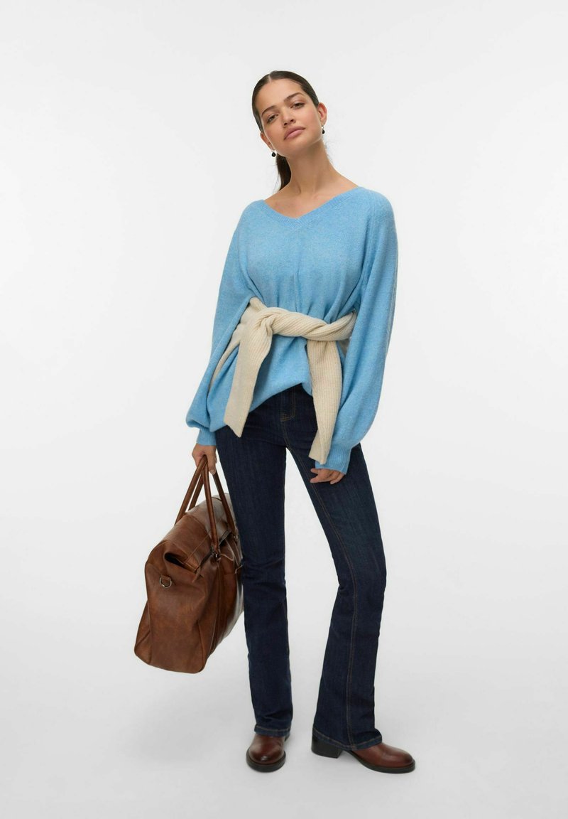 Pull bleu clair avec un col en V et des manches larges, maintenu à la taille par une ceinture tricotée de couleur crème. Jean évasé bleu foncé et sac à main marron.