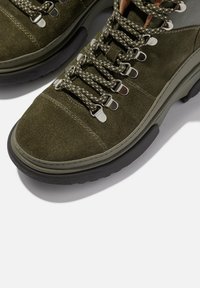 Olijfgroene suède schoenen met ronde veters, metalen oogjes en een gestructureerde zwarte rubberen zool. Met stikaccenten en een ronde neus.