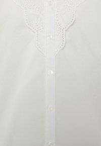 Camisa branca de botão com um delicado acabamento de renda branca ao longo do colarinho, com um padrão de recortes, e botões de plástico transparentes na frente.