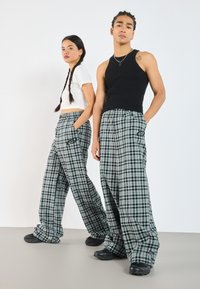 Weite Karo-Hosen in Grün-, Schwarz- und Blautönen mit einem schwarzen Logo-Detail. Kombiniert mit einem weißen Cropped-Top und einem schwarzen Tanktop.