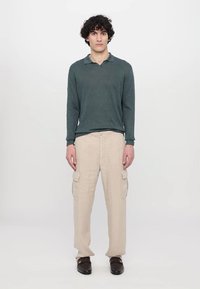 Donker groene polo met lange mouwen, beige cargobroek met zijzakken en donkere schoenen, staand voor een simpele witte achtergrond.