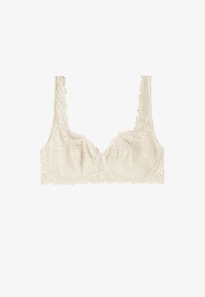 Soutien-gorge en dentelle beige, présentant un motif à carreaux avec des accents floraux. Texture douce avec de larges bretelles en dentelle et sans armature. Design à bonnets ouverts.