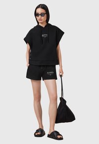 Svart oversized hoodie med snörning, tillsammans med svarta shorts med sidoknappar. Modellen bär en svart tote-väska och har på sig slidesandaler.