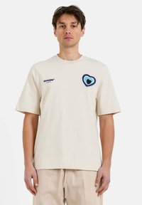 Camiseta de algodón beige con mangas cortas, que presenta un corazón multicolor bordado en el pecho y un sencillo cuello redondo.