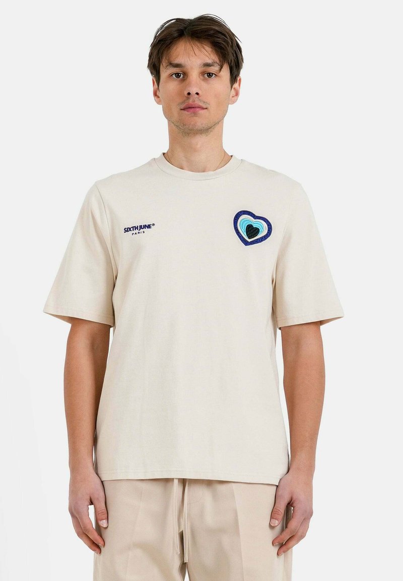 Camiseta de algodón beige con mangas cortas, que presenta un corazón multicolor bordado en el pecho y un sencillo cuello redondo.
