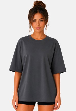 Jeune femme aux cheveux bruns attachés en chignon porte un t-shirt gris foncé oversize et un short noir, debout devant un fond clair uni.