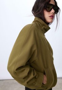 Olivegrüne zip-up Jacke mit hohem Kragen, im lockeren Schnitt, mit seitlichen Taschen und glatter Stofftextur. Mit gesteppten Details akzentuiert.
