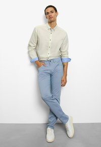 Hellbeiges Hemd mit Knöpfen und blauen Manschetten, kombiniert mit hellblauen Slim-Fit-Chinos und weißen Turnschuhen. Das Model steht mit einer Hand in der Tasche.