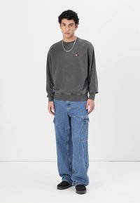 Felpa grigia con polsini e orlo a coste, caratterizzata da un piccolo logo colorato applicato sul petto. Jeans cargo blu a vestibilità ampia con tasche laterali.