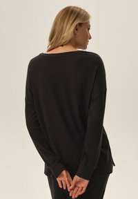 Czarna bluzka z długim rękawem o luźnym kroju, szerokim dekolcie, bocznymi rozcięciami i teksturowanym materiałem, widziana z tyłu.