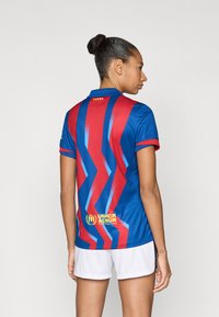 Φανέλα της Μπαρτσελόνα με οριζόντιες μπλε και κόκκινες ρίγες σε μοτίβο ζιγκ-ζαγκ, κοντά μανίκια και λογότυπο του Υπουργείου Ύπατης Αρμοστείας του ΟΗΕ για τους Πρόσφυγες (UNHCR) στο κάτω μέρος της πλάτης.