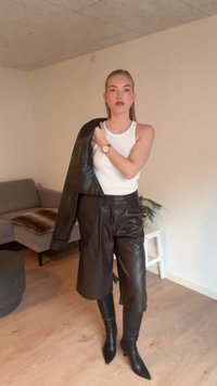 Giacca in pelle marrone drappeggiata sopra una canotta bianca, abbinata a pantaloncini in pelle marrone e stivali neri al ginocchio con punta affilata. Design semplice e minimalista.