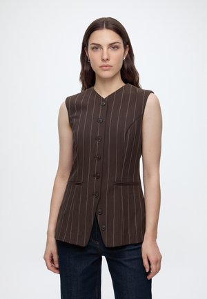 Donna con lunghi capelli castani che indossa un gilet marrone a righe senza maniche con bottoni e jeans blu scuro su sfondo bianco.
