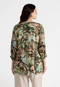 Blumenbluse in einer grünen und beigen Farbpalette aus leichtem Material, mit 3/4-Ärmeln und bequemer Passform sowie Faltenlegung auf der Rückseite.