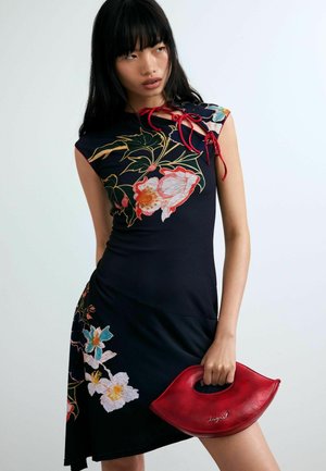 Mujer vestida con un vestido floral negro con detalles de lazo rojo, sosteniendo un bolso rojo en forma de labios sobre un fondo claro y liso.