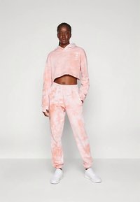 Roza kaklasa krāsainā kapučveida džemperis ar garām piedurknēm un elastīga vidukļa sporta bikses, kopā ar baltām kedām. Audums izskatās mīksts un ar tekstūru.